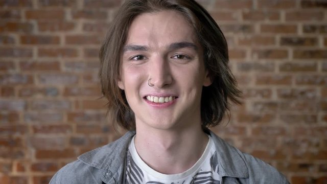 Young long hair man with nose ring is watching at camera, giggling, brick background