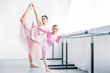 beautiful adult and little ballerinas stretching and smiling at camera in ballet school
