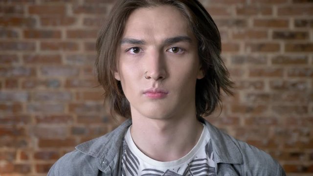 Young serious long hair man with nose ring is watching at camera, smiling, brick background