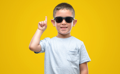 Dark haired little child wearing sunglasses surprised with an idea or question pointing finger with...