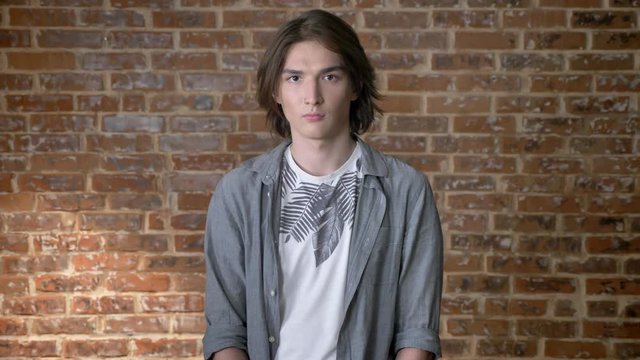 Young serious long hair man with nose ring is watching at camera, brick background