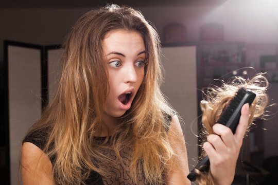 Shocked young pretty brown haired girl standing in a beauty salon discovers a big amount of lost hair on the hair brush