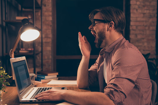 Portrait of attractive, tired, drowsy man in shirt with stubble cover his wide open mouth with palm, yawn, sitting in workplace, workstation, working at night time
