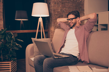 Obraz premium Portrait of cheerful, positive, domestic guy in denim outfit, jeans, shirt, holding arms behind the head, watching film, looking at screen of computer, having relax, fun, pause during work at night