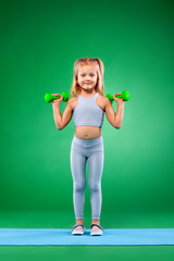 Kid girl doing fitness or yoga exercises with dumbbells isolated on green background