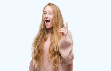 Blonde teenager woman wearing pink sweater pointing finger up with successful idea. Exited and...