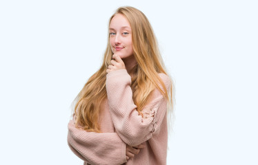 Blonde teenager woman wearing pink sweater looking confident at the camera with smile with crossed arms and hand raised on chin. Thinking positive.