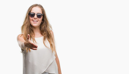 Young blonde woman wearing sunglasses smiling friendly offering handshake as greeting and welcoming. Successful business.