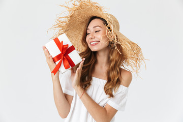 Young beautiful woman 20s wearing big straw hat smiling and holding present box with red ribbon, isolated over white background