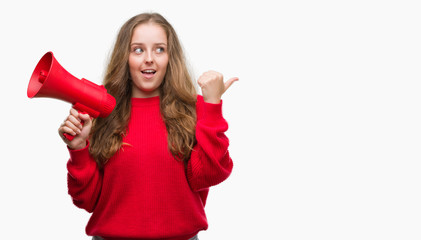 Young blonde woman holding red megaphone pointing and showing with thumb up to the side with happy...