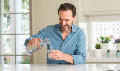 Middle age man drinking a glass of water with a happy face standing and smiling with a confident...