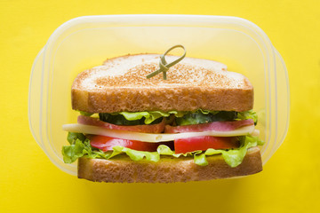 Sandwich with cheese, ham and fresh vegetables in a container on a yellow bright background.