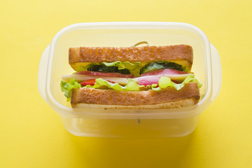Sandwich with cheese, ham and fresh vegetables in a container on a yellow bright background.
