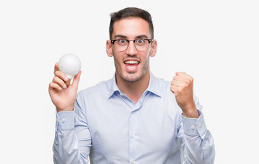 Handsome young man holding lightbulb as idea screaming proud and celebrating victory and success very excited, cheering emotion