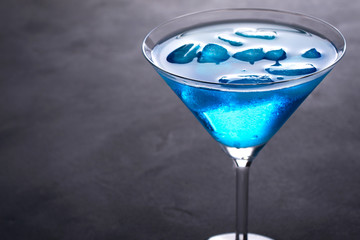 Blue cold cocktail in martini glass with ice and clear dew drops on  glass on a gray concrete background with space for text