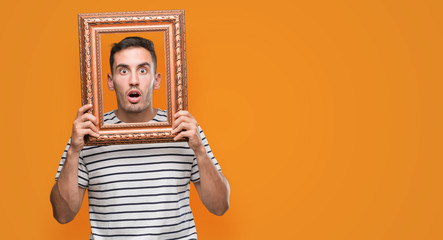 Handsome young man looking through vintage art frame scared in shock with a surprise face, afraid and excited with fear expression