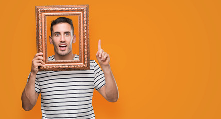 Handsome young man looking through vintage art frame surprised with an idea or question pointing...