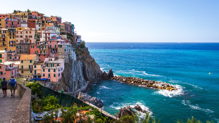 A couple walks along a brick path in Manarola, Cinque Terre, Italy. Shown are the colorful houses on the hills and the Tyrrhenian sea in the background