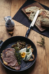 A view of a grilled beefsteak and an egg in a pan surrounded by eggs, garlic and olive oil