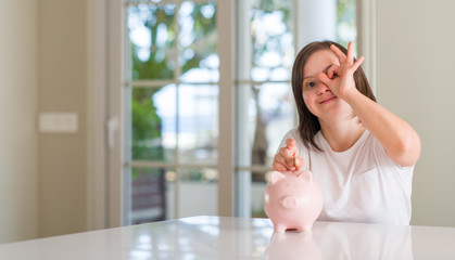 Down syndrome woman at home holding piggy bank with happy face smiling doing ok sign with hand on eye looking through fingers