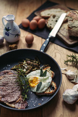 A view of a grilled beefsteak and an egg in a pan surrounded by eggs, garlic and olive oil