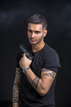 Young handsome man holding a hand gun, wearing black t-shirt, on black background in studio