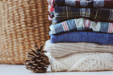 fall casual woman fashion clothes set. Stack of plaid shirts and knitted sweaters with pine cone and basket on background. Cozy stylish womanswear.