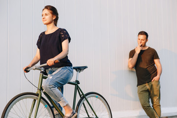Gorgeous young girl riding a bicycle, while stylish handsome guy smoking and looking with admiration at her. Outdoors.