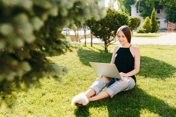 Young student girl in casual clothes working on a laptop, studying, preparing for exams, while sitting on the grass, in the park.