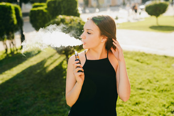 Fototapeta premium Side view of young beautiful girl vaping e-cigarette, outdoors. Wearing black stylish singlet.