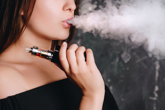 Close-up view of charming young woman with short hair smoking an e-cigarette, cloud of vapor.