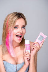 excited young woman with colored bob cut holding hair care supplies and looking at camera isolated on grey