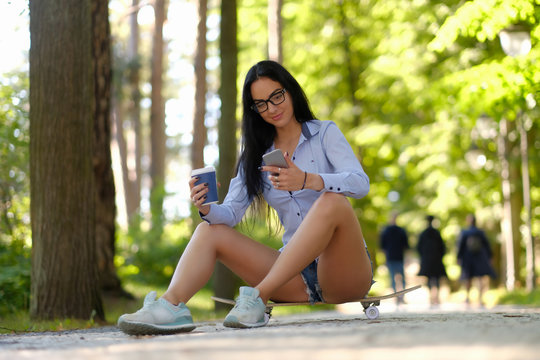Gorgeous sexy brunette girl in glasses wearing a shirt and shorts sitting on a skateboard and holds cup of takeaway coffee and using a smartphone at the park.