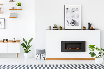 Poster above black fireplace in white living room interior with plants and grey stool. Real photo