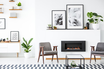 Fireplace between grey armchairs in modern living room interior with posters and plants. Real photo