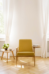 Yellow wooden armchair next to table with plant in white living room interior with drapes. Real photo