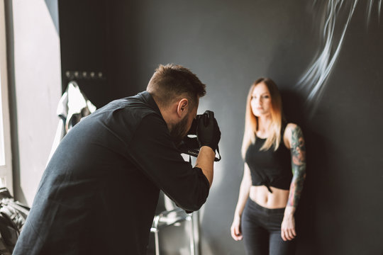 Tattoo master taking photos on camera of tattooed girl standing on black background in modern studio