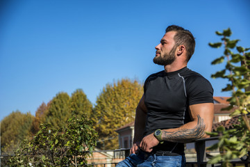 Handsome dark haired young man standing in a balcony, looking out and down at the view,wearing a black T-shirt while, in summer