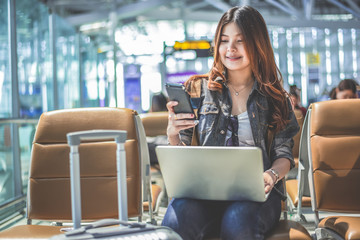 Young Asian female passenger using laptop and smart phone while sitting on seat in terminal hall and waiting for flight in airport . People lifestyles and Happy woman. Technology and Travel concept.