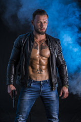 Handsome man wearing leather jacket on naked muscular torso holding hand gun, on dark smoky...