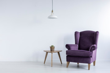 A wooden table and a fancy, dark purple, wing armchair in an empty living room interior with white walls and place for furniture. Real photo.