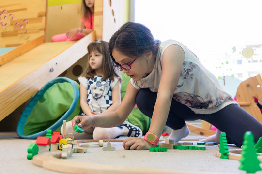 Cute girl with a serious facial expression playing with a wooden train circuit on the floor, during free playtime in the classroom of a modern kindergarten