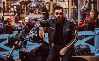 Fototapeta na wymiar Stylish fashionable biker in sunglasses dressed in a black leather jacket, sitting on his custom-made retro motorcycle near terrace of cafe.