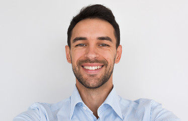 Closeup portrait of happy handsome young male smiling with healhty toothy smile looking at the camera, making self portrait. Cheerful unshaven man wearing blue shirt posing in studio background.People