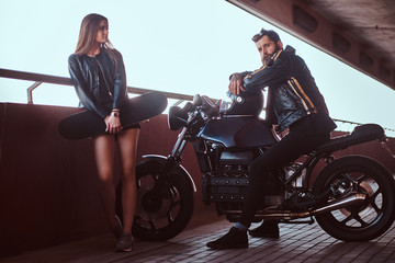 Fototapeta na wymiar Portrait of an attractive couple - brutal bearded biker in black leather jacket sitting on a motorcycle and his young sensual brunette girlfriend holds skateboard.