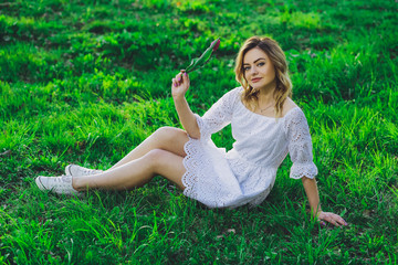 Young beautiful blonde girl in white dress with a flower tulip on a background of green grass