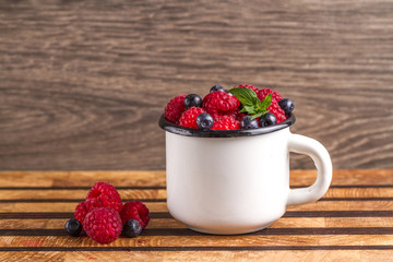 Berries of raspberries, blueberries and a sheet of fresh mint in a white cup on a wooden background.