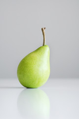 One green yellow pear with a twig stands on a white table, a reflection, a bright light, minimalism and free space for text.