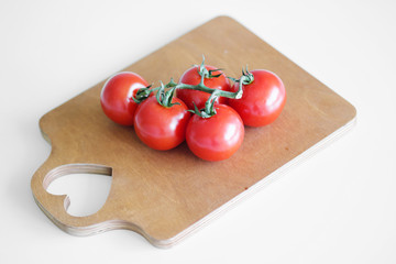 Cherry tomatoes on a branch lie on a wooden cutting board on a white table. The heart is cut out on the board.
