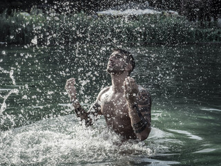 Handsome, hot young bodybuilder in the sea, splashing water up, showing his muscular torso and arms